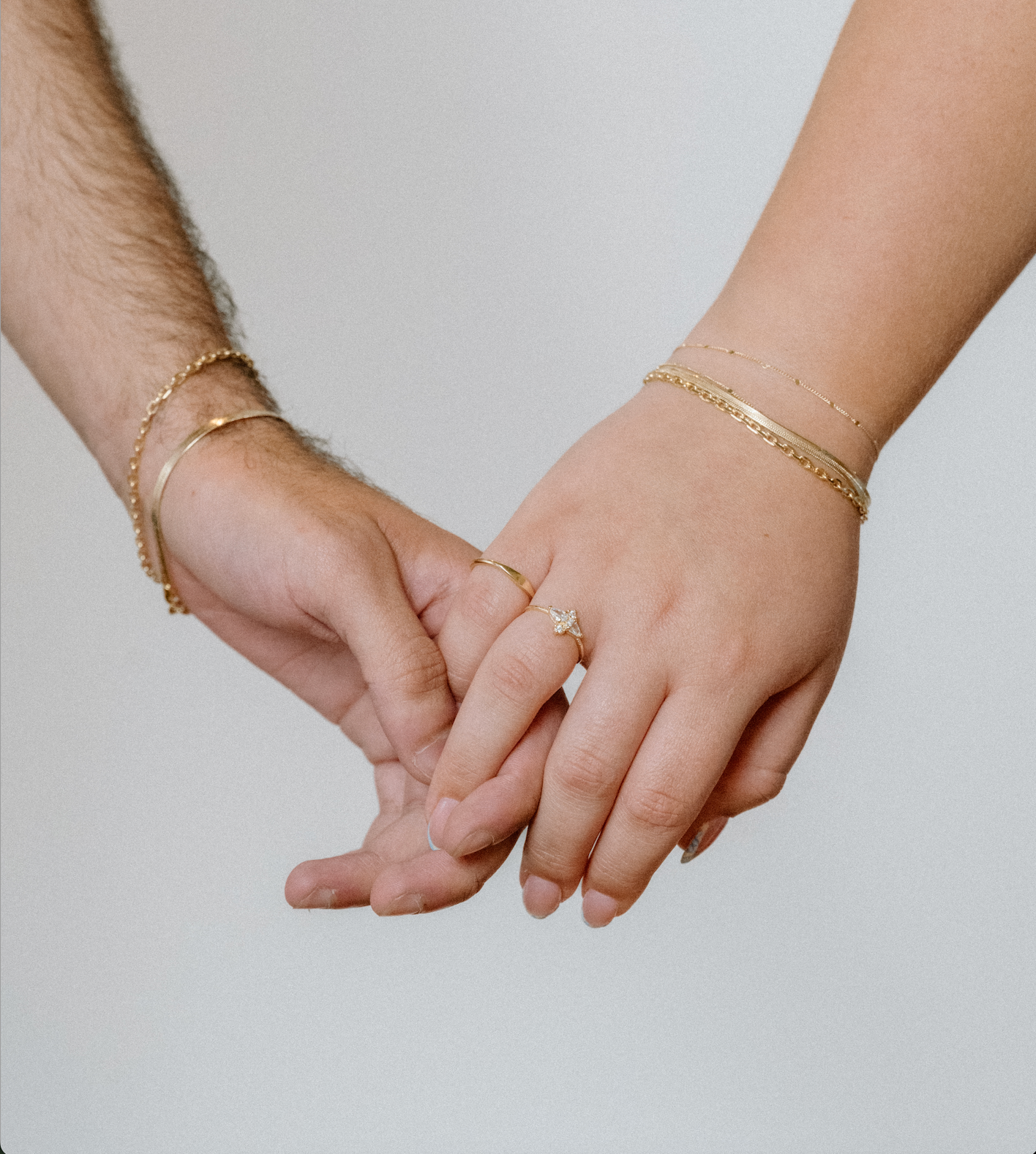 Two hands gently holding, one wearing a pavé butterfly ring, gold herringbone bracelet, and gold chain bracelet, the other wearing a gold herringbone bracelet and gold curb chain bracelet.