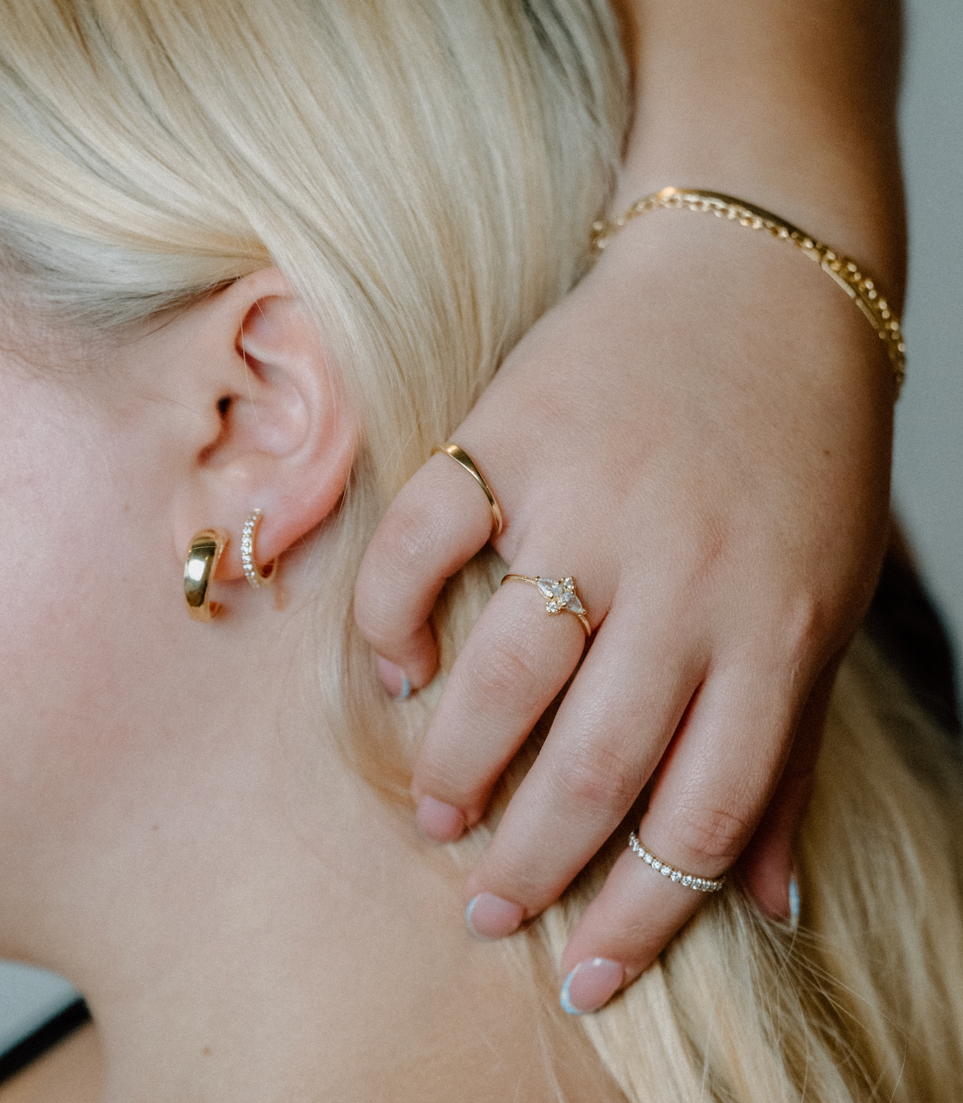 A woman's ear and hand styled with gold hoop earrings, a pavé diamond ring, and stackable gold bands in a layered jewelry look.