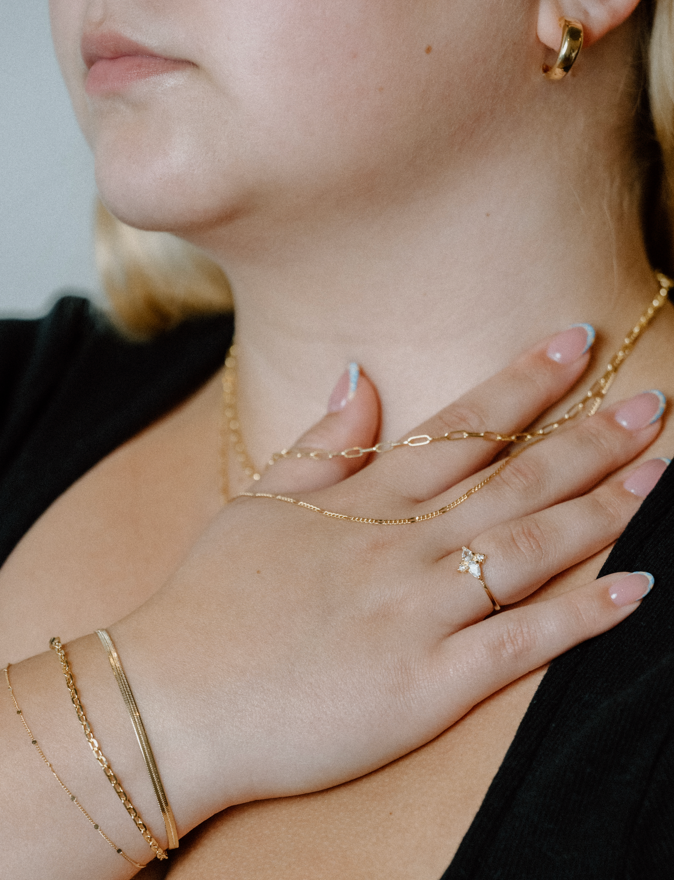A woman wearing layered gold chain necklaces, minimalist bangles, and a pavé ring with hand over chest.