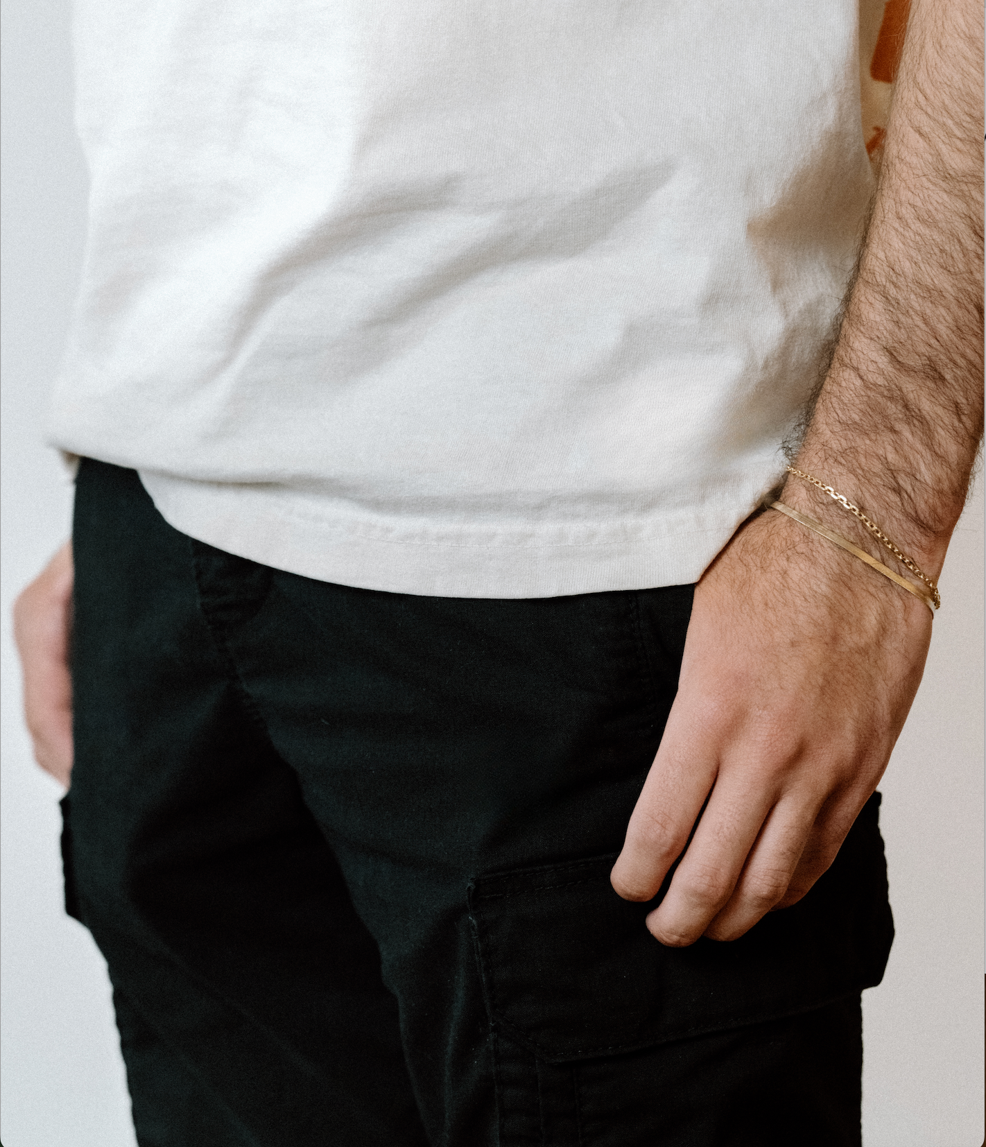 Man wearing a gold herringbone bracelet and gold chain bracelet with a white t-shirt and black cargo pants, hand resting casually by his side.