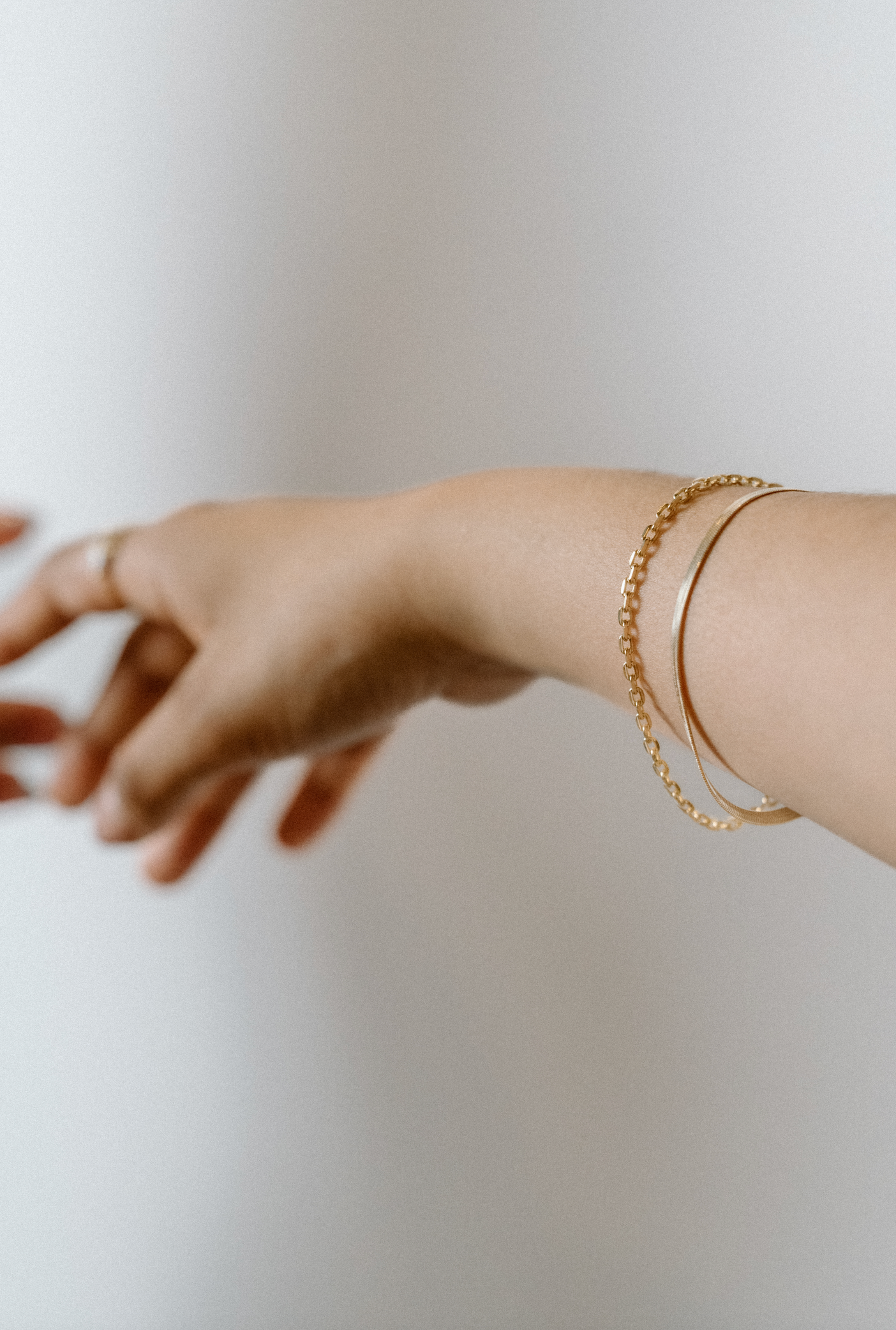 Extended arm wearing a gold chain bracelet and herringbone gold bracelet, shown in soft motion against a neutral background.